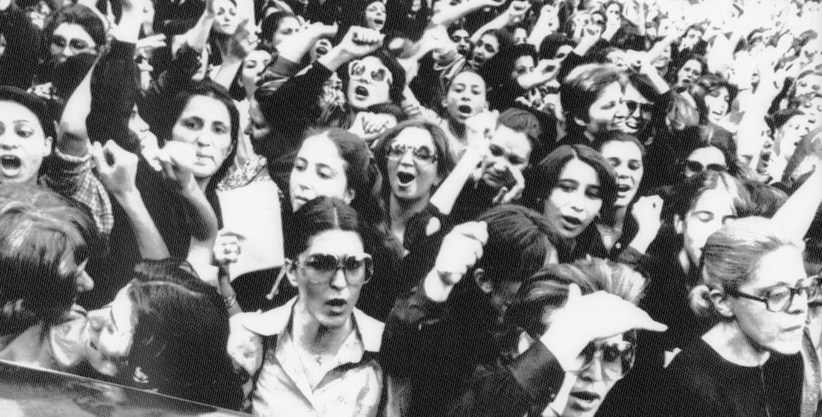 An old black-and-white photo of women protesting in Iran in the 80s. Many of them are yelling, with their fists in the air. T