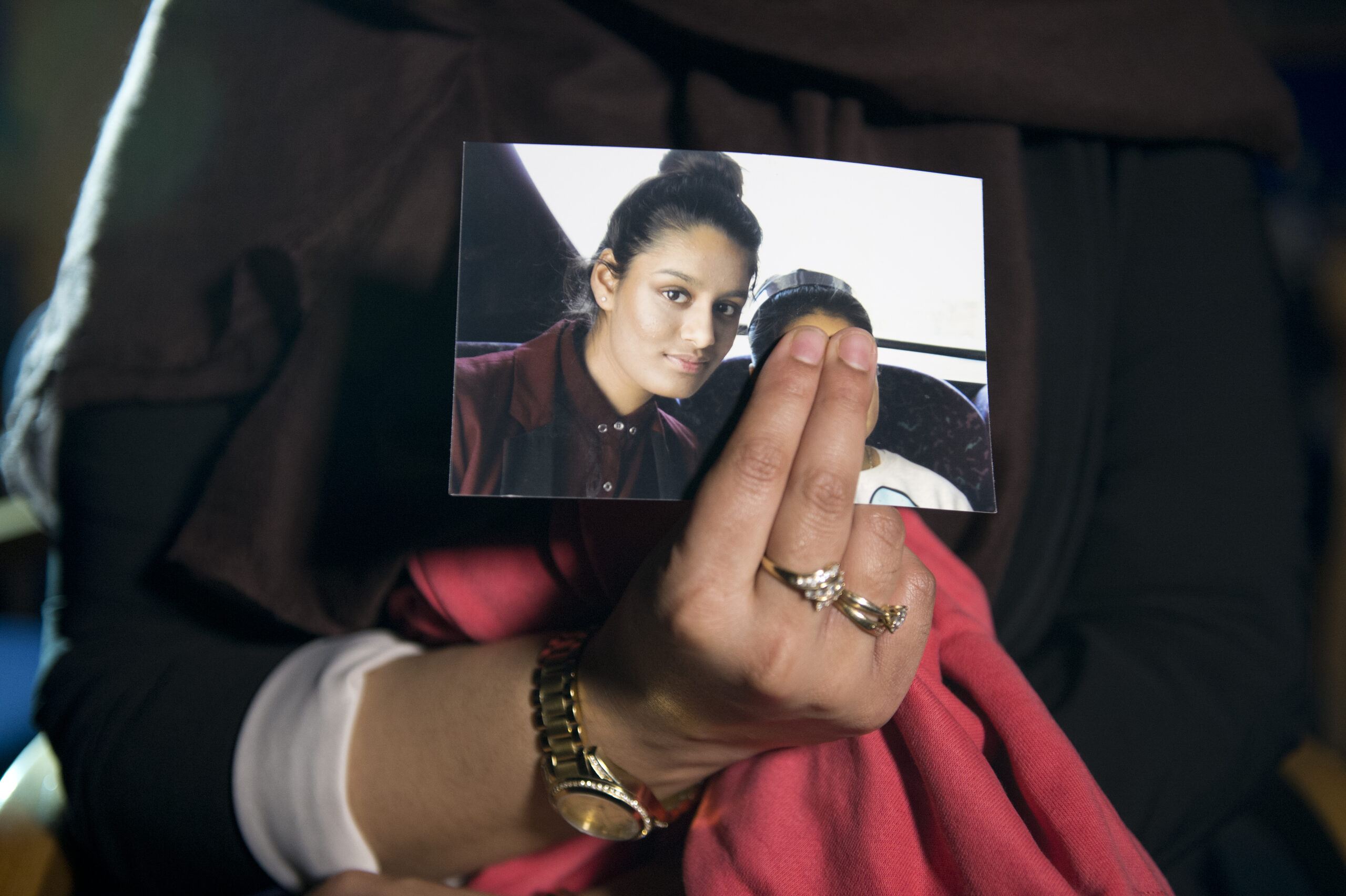 The older sister of Shamima Begum, Renu Begum, holds a photo of her sister with a child. Two of her fingers obscure the child's face. She is wearing rings on two of her fingers and a watch on her wrist. In the photo, Shamima's hair is tied in a bun and she's wearing a burgundy button down shirt buttoned to the top, and a matching blazer over it. She is looking directly at the camera.