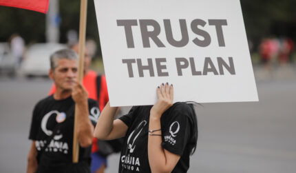 Woman in QAnon shirt holding sign "Trust the plan"