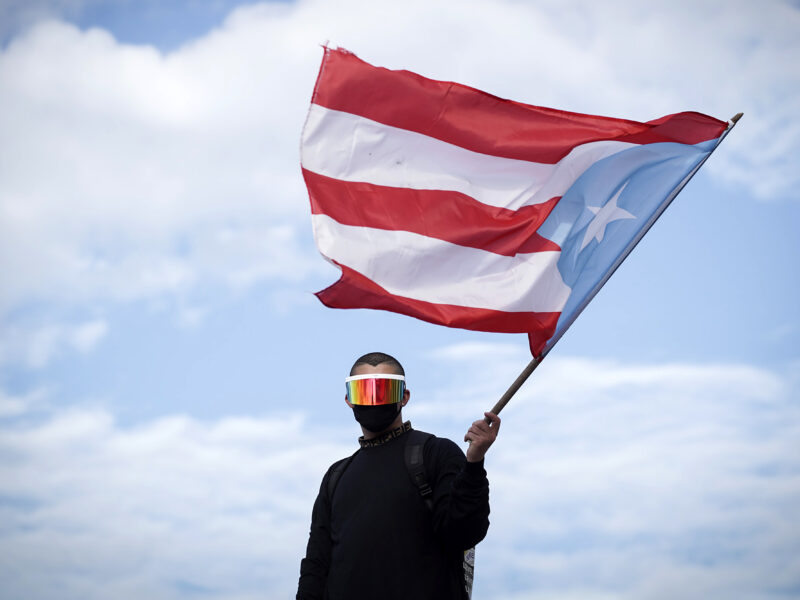 Bad Bunny wearing a black long sleeved shirt, a black cloth face mask, and giant sunglasses that shield his eyes, looking directly at the camera while waving a giant Puerto Rican flag.
