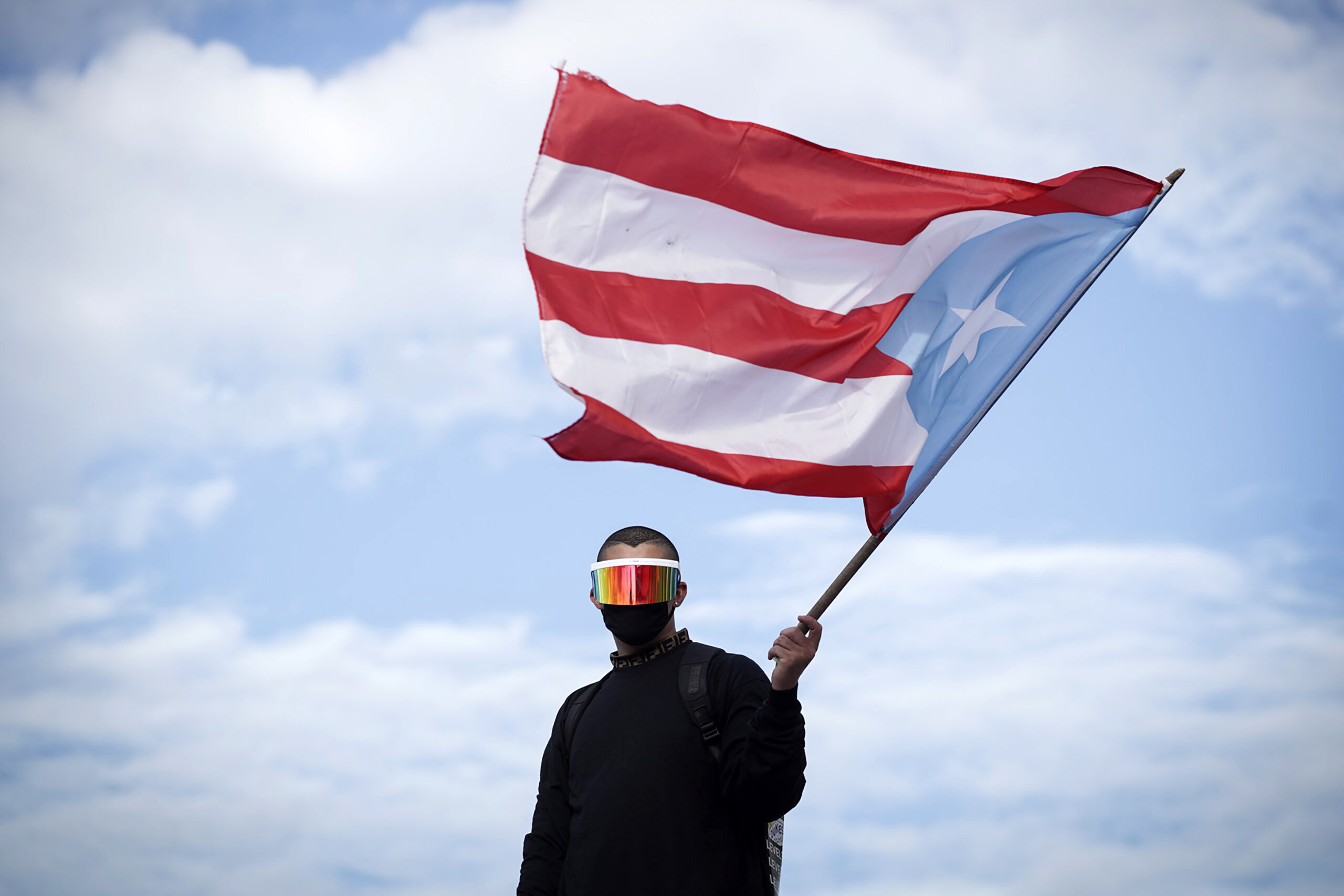 Bad Bunny wearing a black long sleeved shirt, a black cloth face mask, and giant sunglasses that shield his eyes, looking directly at the camera while waving a giant Puerto Rican flag.