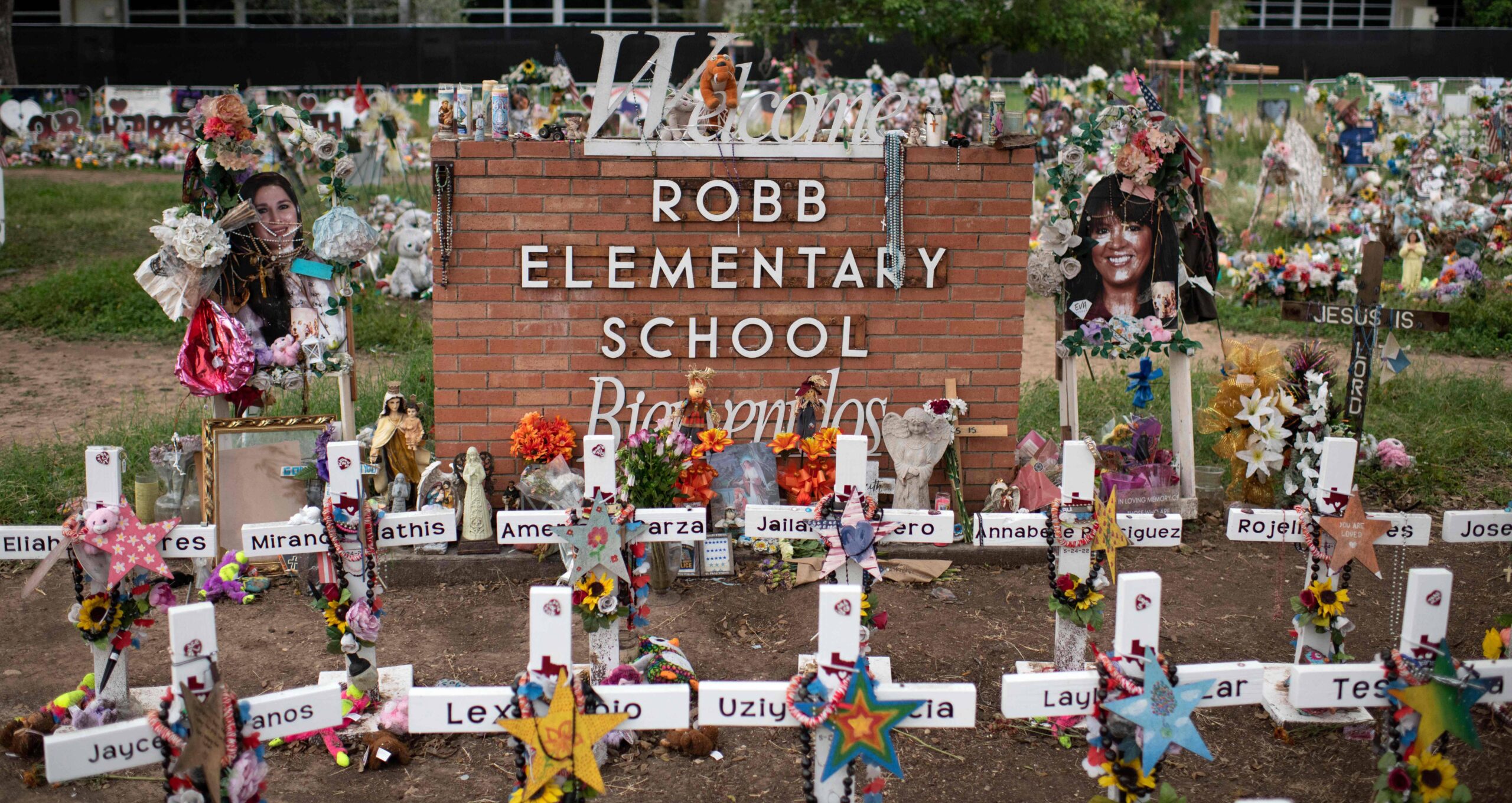 A brick welcome sign for Robb Elementary School. All around it are white crosses with the victims' names written on them, flowers, candles, and toys