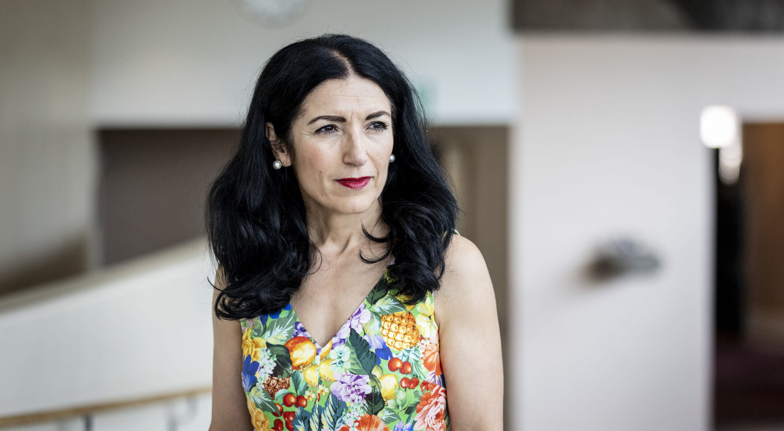 Photograph of Kurdish exile and former Swedish Parliament member Amineh Kakabaveh, a woman in her early 50s with black wavy hair below her shoulders. She's wearing red lipstick and pearl earrings, and a colorful dress with a pattern of various fruits and flowers. She is looking off to the right.