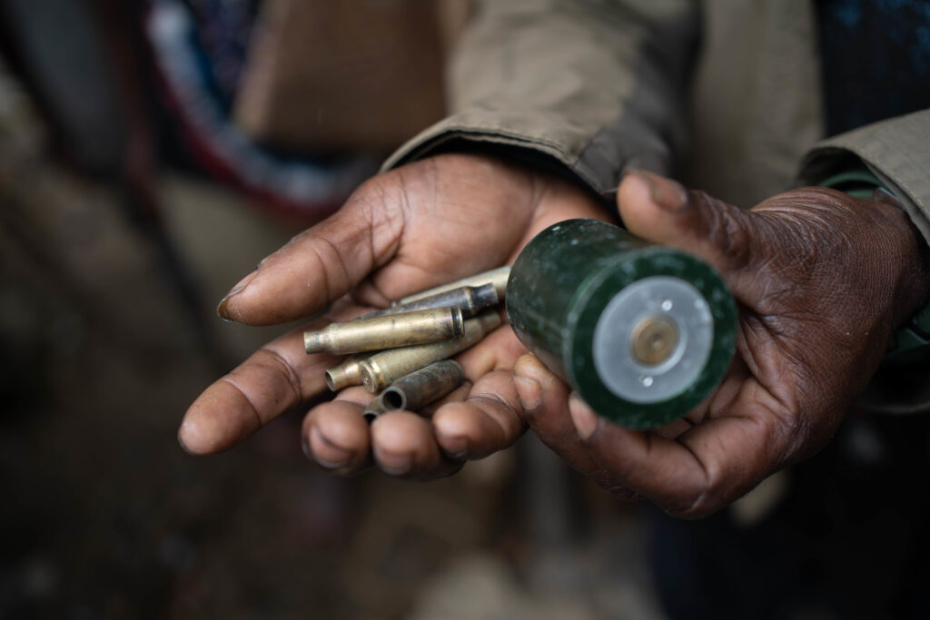 A close-up photograph of hands holding recovered ammunition from an aerial attack in West Papua, including what appear to be large brass-colored shells and a larger canister. The only thing in focus are the hands and the weaponry, but you can see the person holding the shells is wearing a dark gray-green jacket. 