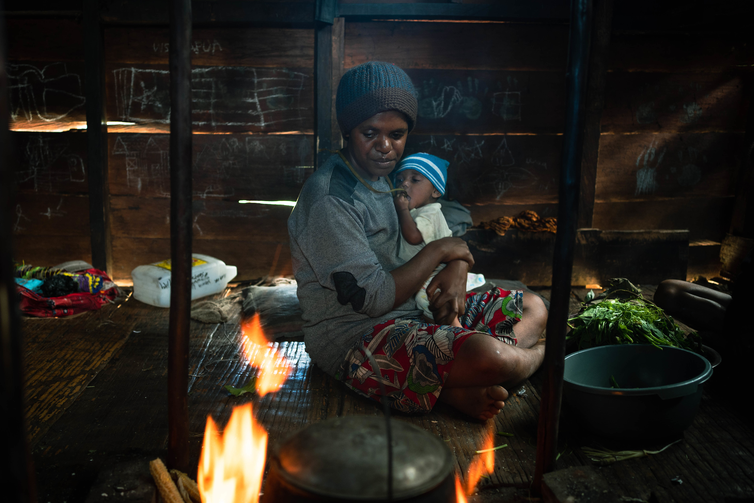 A mother sits cross-legged inside of a dark wooden structure, looking at a pot on a fire. She is holding a young child, who is grasping her necklace and putting it in their mouth. The woman is wearing a dark blue and black knit cap, a long-sleeved gray shirt, and a red skirt patterned with large leaves. The child is wearing a lighter blue knit hat and a white garment. There are chalk drawings on the wooden walls behind them.