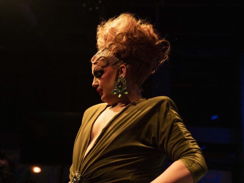 Drag queen Vanity is under a spotlight in the foreground, while most of the background is black. Her red hair is teased high in an up-do, with the front smoothed back. She's in profile, looking off to the side, and is wearing a large earring with many large gemstones. She's wearing a low cut muted green dress, with a crystal broach at the waist.