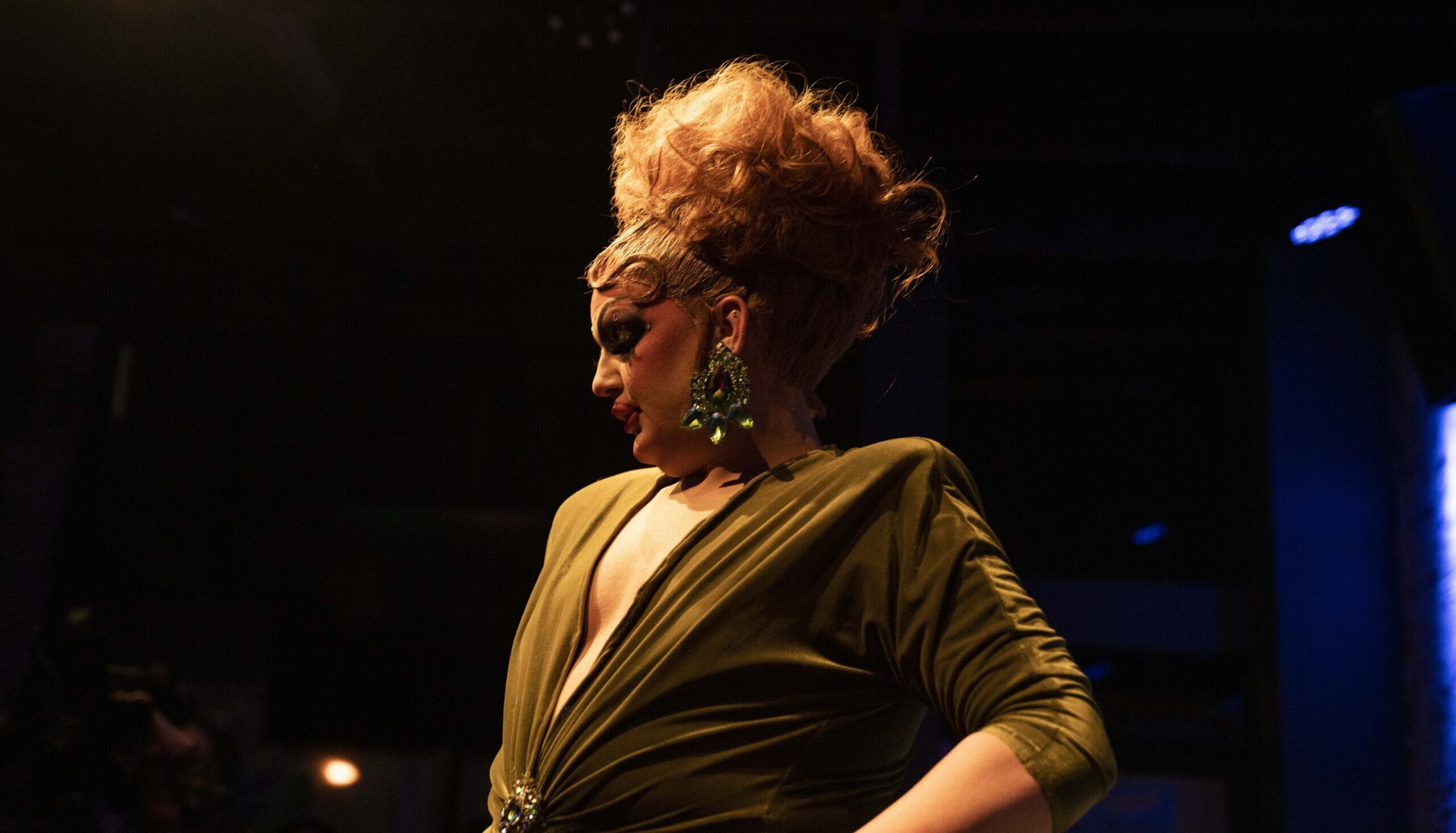 Drag queen Vanity is under a spotlight in the foreground, while most of the background is black. Her red hair is teased high in an up-do, with the front smoothed back. She's in profile, looking off to the side, and is wearing a large earring with many large gemstones. She's wearing a low cut muted green dress, with a crystal broach at the waist.