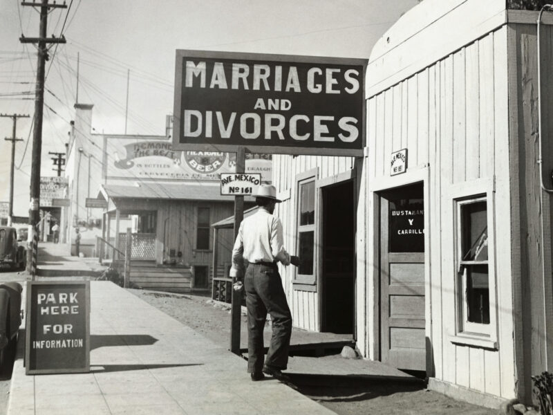A vintage, black-and-white photo of a man walking into a building with a large "MARRIAGES AND DIVORCES" sign. He's wearing a long sleeved white button-down, high-waisted pants, and a cowboy hat. You can't see his face. On the site, there's a sandwich board sign that says "park here for information," next to an old-fashioned car.