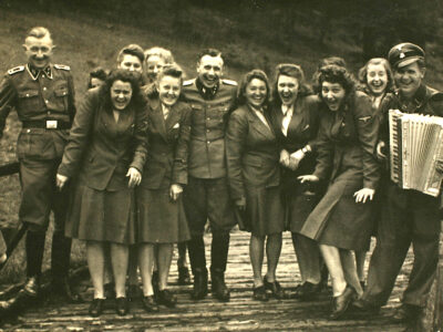 A sepia photo from 1944 of a group of about a dozen Nazi officers and auxiliaries—including Karl Höcker in the center—all openly laughing. The photo was taken at Solahütte, a "resort" where Nazis who worked at Auschwitz would vacation on weekends. They are all in uniform: The women wear long sleeved blazers and knee length skirts with flat shoes, and the men wear long sleeved military jackets and trousers tucked into tall boots. The man on the right is wearing a Nazi hat and is holding an accordion. They're standing on what appears to be a wooden bridge. There is grass behind them.