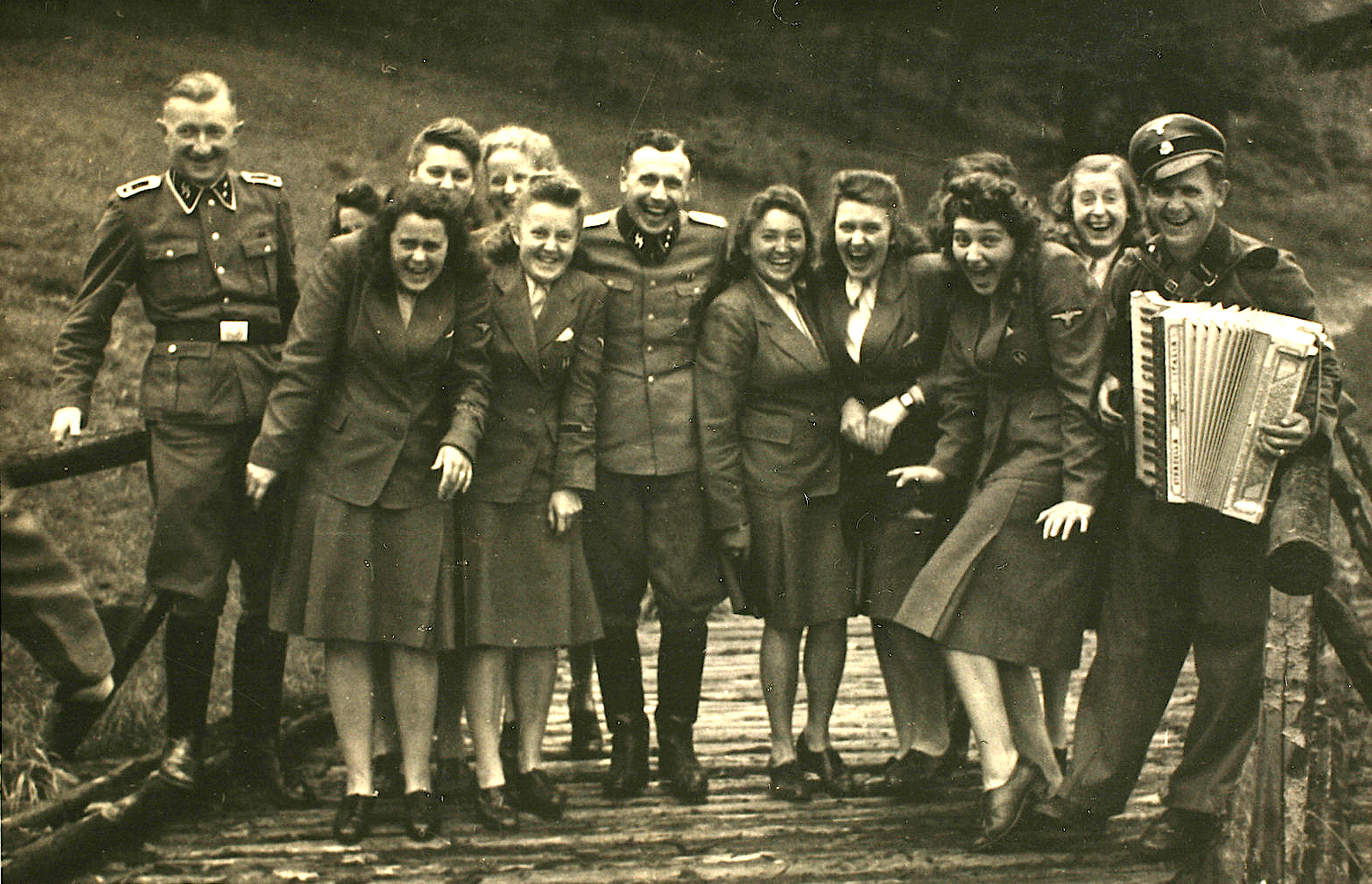 A sepia photo from 1944 of a group of about a dozen Nazi officers and auxiliaries—including Karl Höcker in the center—all openly laughing. The photo was taken at Solahütte, a "resort" where Nazis who worked at Auschwitz would vacation on weekends. They are all in uniform: The women wear long sleeved blazers and knee length skirts with flat shoes, and the men wear long sleeved military jackets and trousers tucked into tall boots. The man on the right is wearing a Nazi hat and is holding an accordion. They're standing on what appears to be a wooden bridge. There is grass behind them.