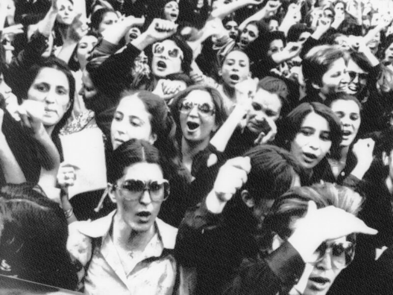 An old black-and-white photo of women protesting in Iran in the 80s. Many of them are yelling, with their fists in the air. They're protesting an (at the time) newly enforced dress code for women in Iran, requiring women to dress a certain way or else lose their jobs.