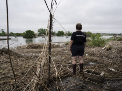 "A resident of Fedorivka is seen standing outside her flooded garden," caused by the collapse of the Kakhovka Dam. She is wearing rainboots, and wearing a black tshirt that says "Espresso Expert" across the back. We do not see her face. She is surrounded by dry debris but just a few feet in front of her we see a good amount of water still from the flooding.