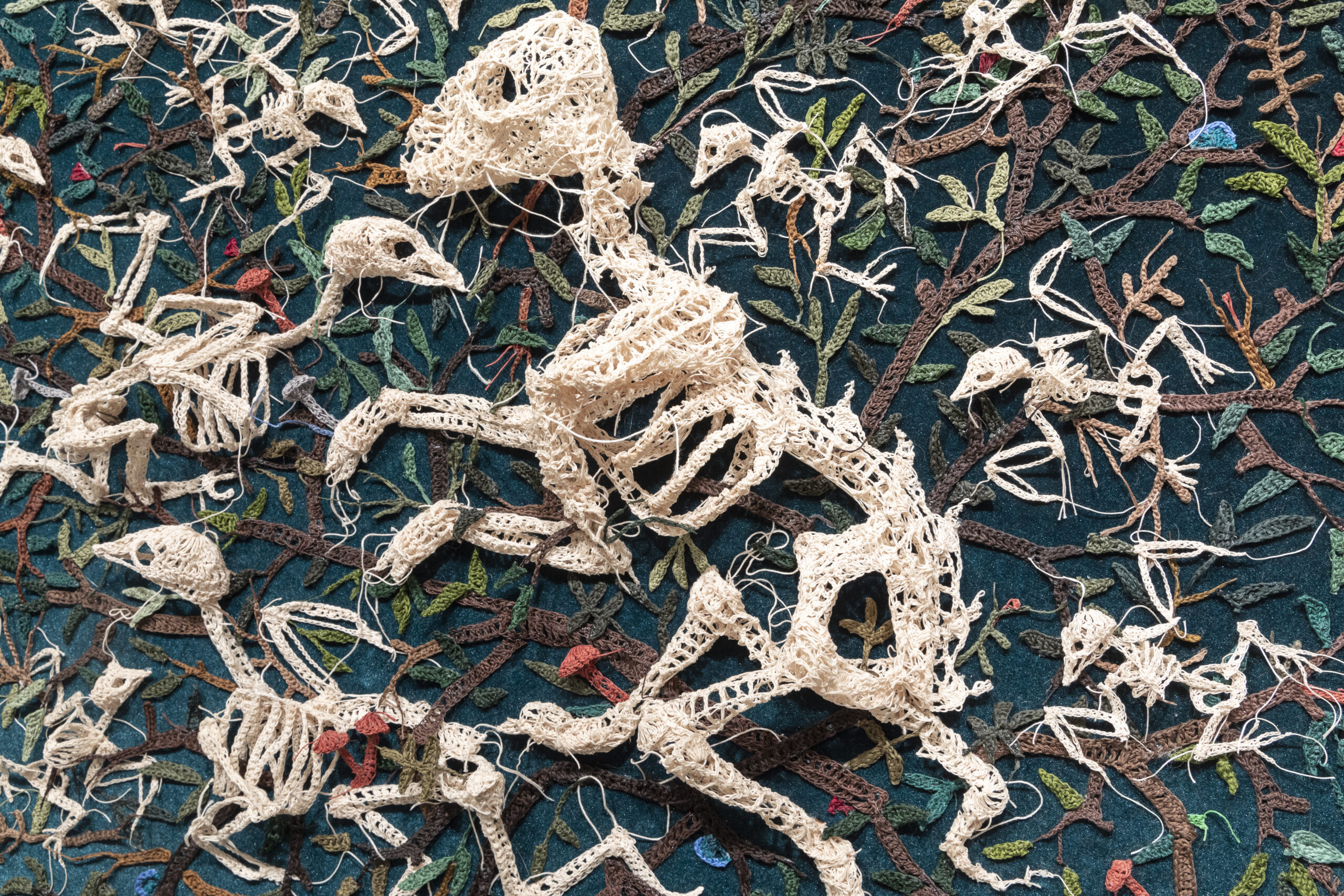 A close up of a piece of embroidery artwork, depicting off-white dinosaur skeletons emerging from and tangled up in a tree.