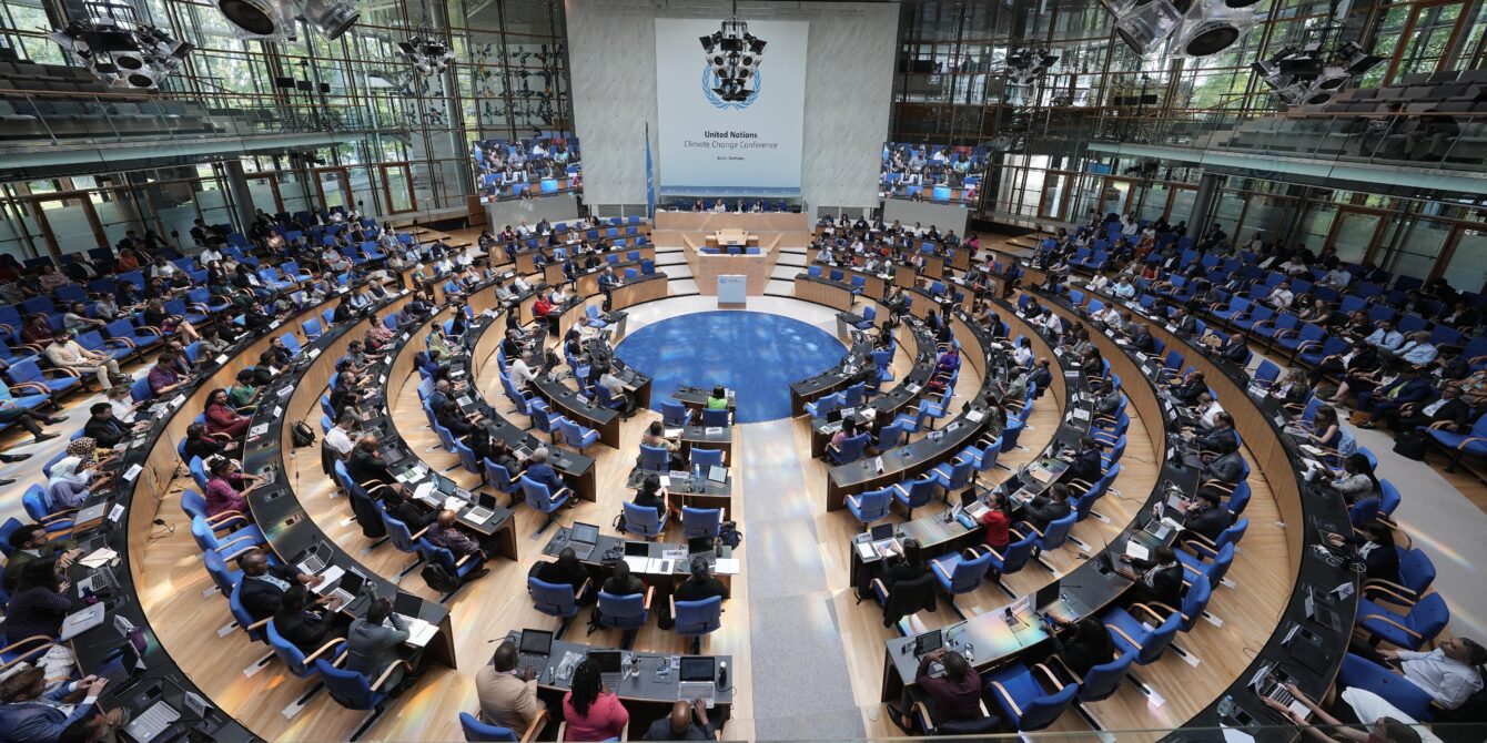 An overhead shot of the climate conference in Bonn last June. Desks are arranged in a circle in a high-ceilinged conference room with floor to ceiling windows. Many of the seats are occupied by representatives from various countries.