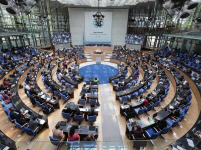 An overhead shot of the climate conference in Bonn last June. Desks are arranged in a circle in a high-ceilinged conference room with floor to ceiling windows. Many of the seats are occupied by representatives from various countries.