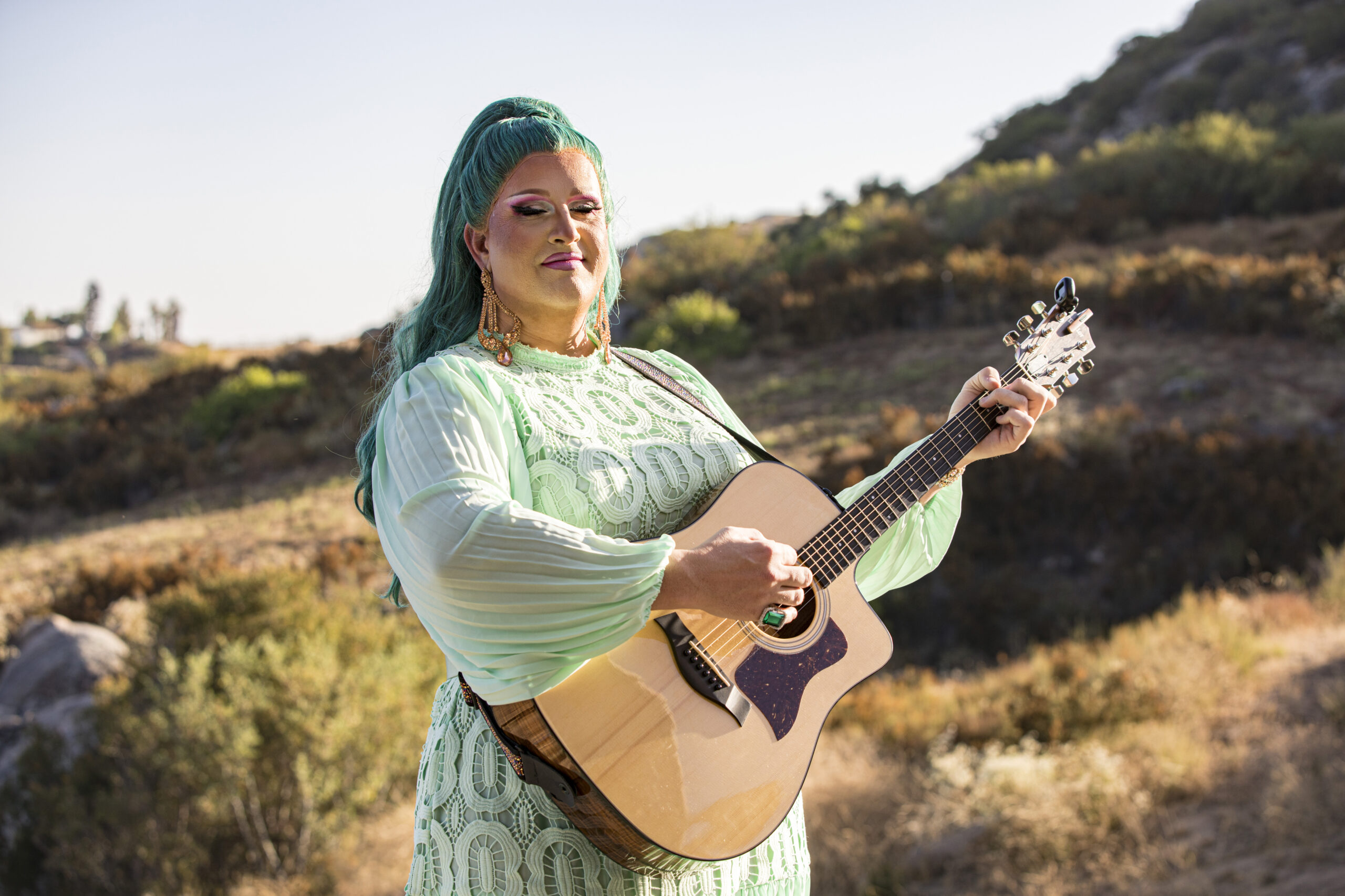 Drag performer Flamy Grant poses prior to the Songbirds of Ramona Ranch show at Ramona Ranch Winery on August 04, 2023 in Ramona, California. She has teal hair in a high ponytail, and is wearing a mint colored long sleeved, sheer dress. She is strumming at an acoustic guitar, her eyes closed.