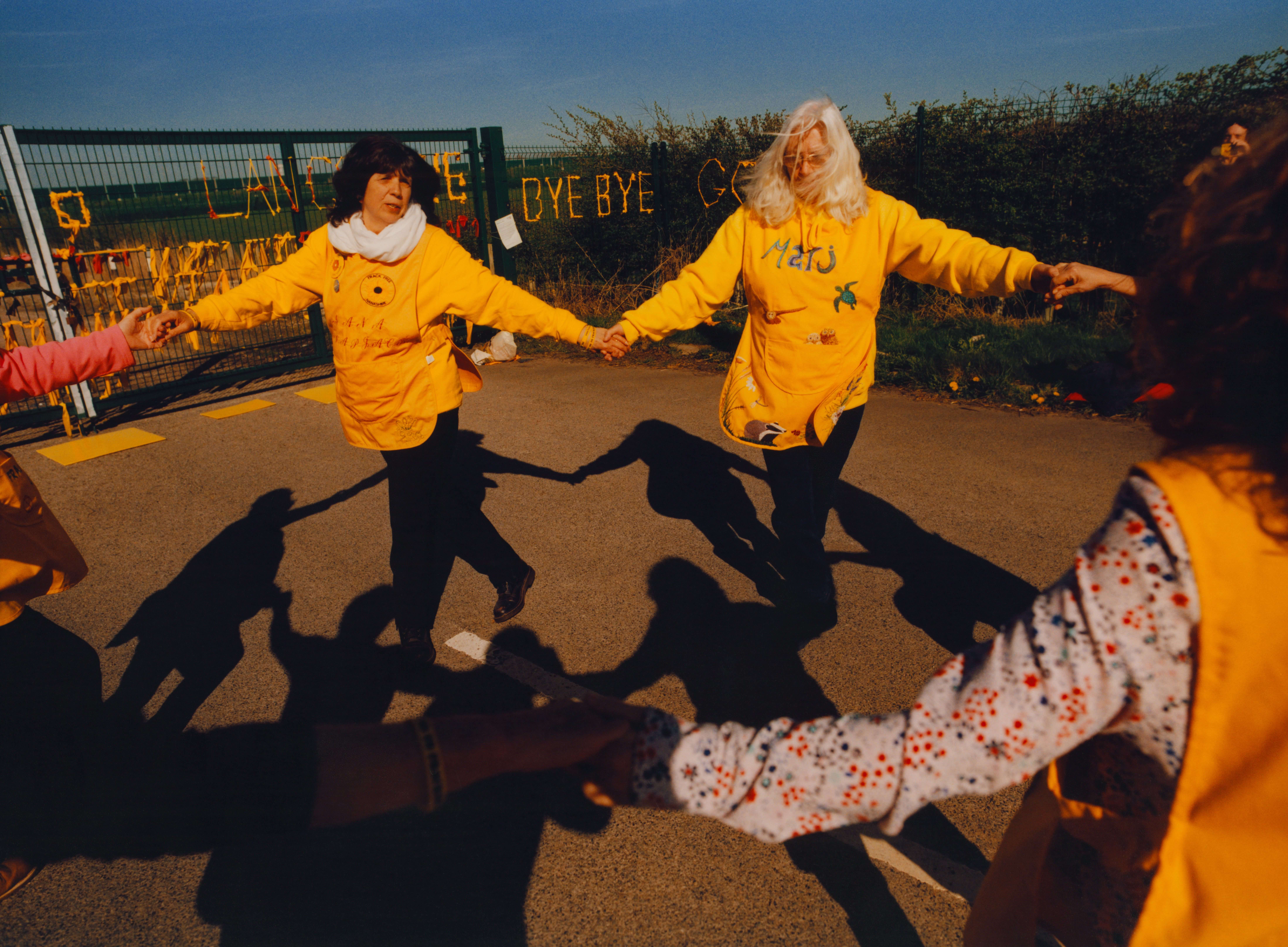 Older women in yellow tabards dance in a circle in front of a former fracking site, holding hands. The photo is reminiscent of Matisse's La Danse. 