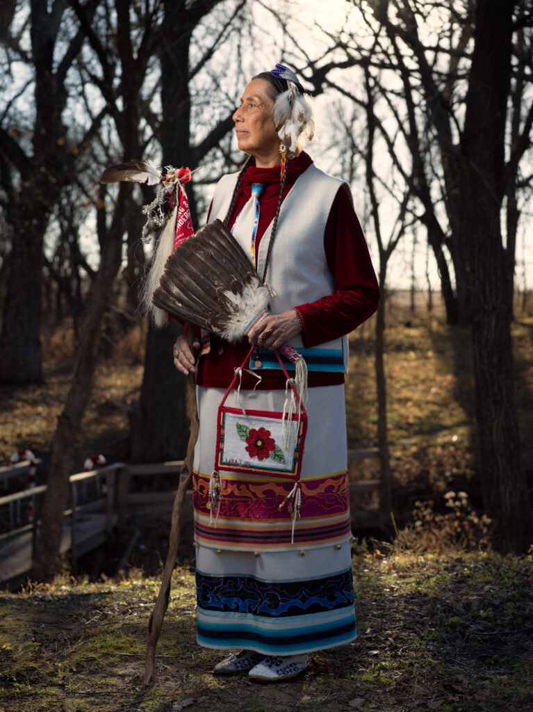 A portrait of Casey Camp-Horinek wearing a feather headpiece and beaded earrings, and holding a small purse with a red flower on it. She's wearing a red velvet turtleneck with a white vest over it, a layered skirt, and beaded shoes. 