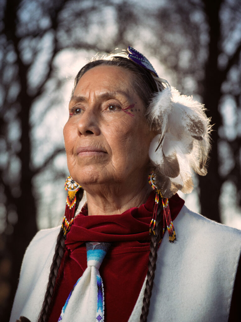 A portrait of Casey Camp-Horinek wearing a feather headpiece and beaded earrings. She's wearing a red velvet turtleneck with a white vest over it.