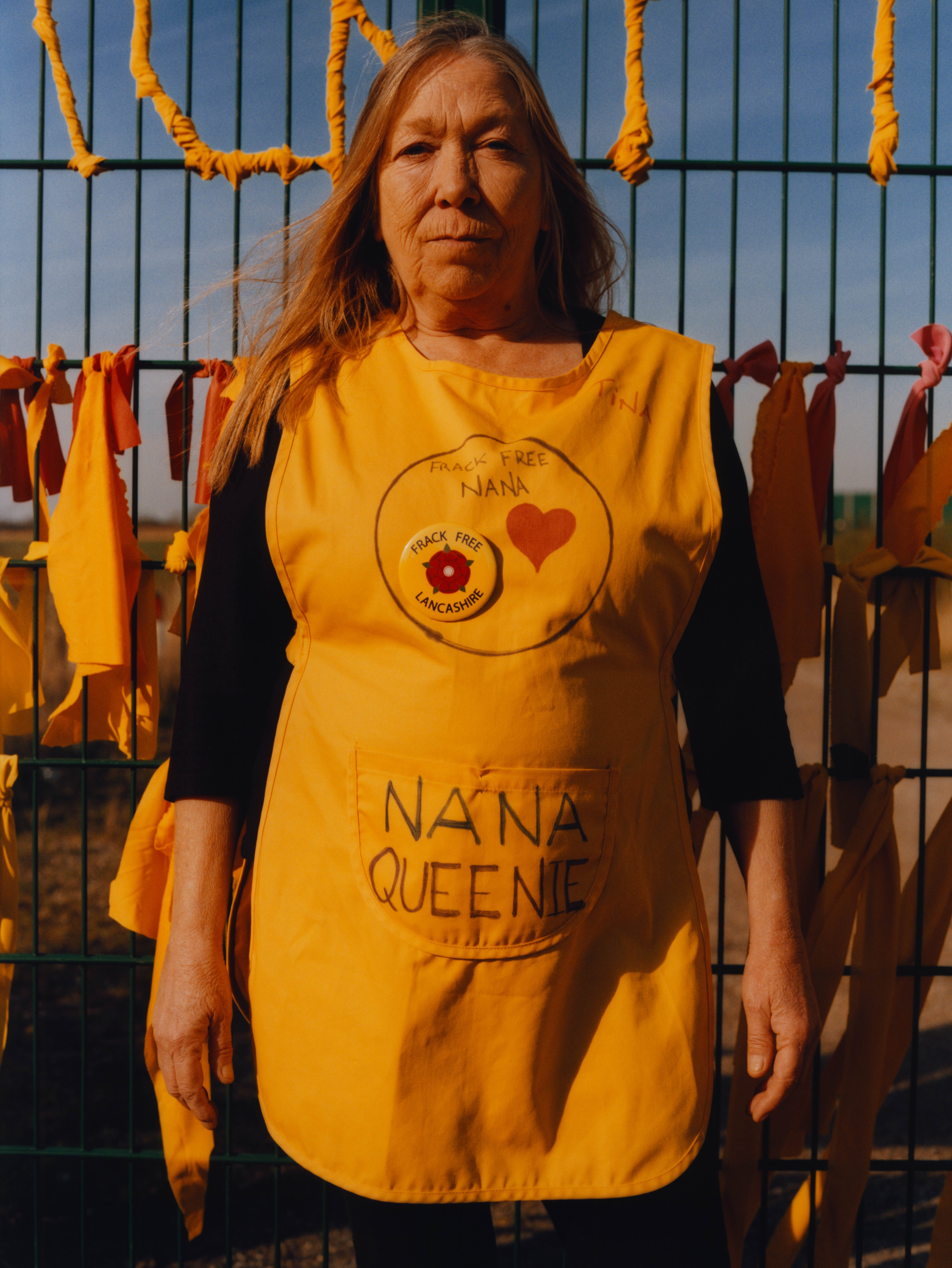 A portrait of Tina Rothery, an older woman with long, straight strawberry blonde hair, wearing a yellow tabard and standing in front of a fence with yellow ribbons tied across it. 