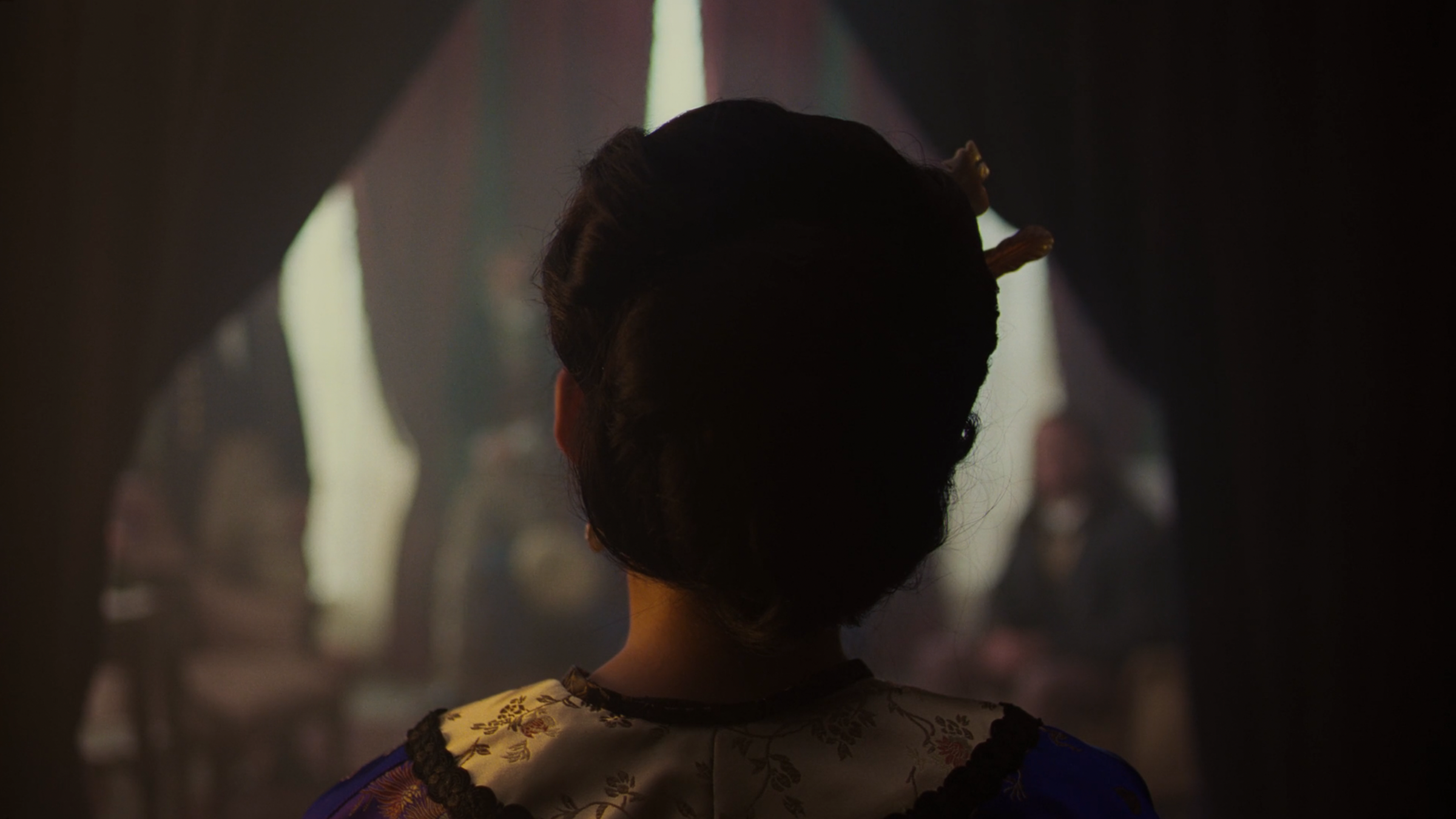 A film still from Astonishing Little Feet, a short film about Afong Moy by Maegan Hoaung. We see the back of the actress playing Moy's head, facing forward, a curtain in front of her drawn to reveal her blurred audience of four men.