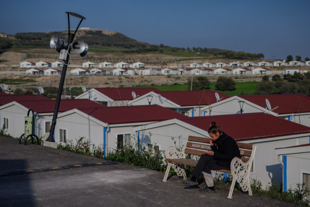 A woman living in a prefabricated city sits on a bench and talks on the phone. She is wearing all black and sandals, her hair tied in the bun. In the background, the homes are all small and light gray with dark red roofs and a line of blue. Some have satellites. Photo: Can Erok