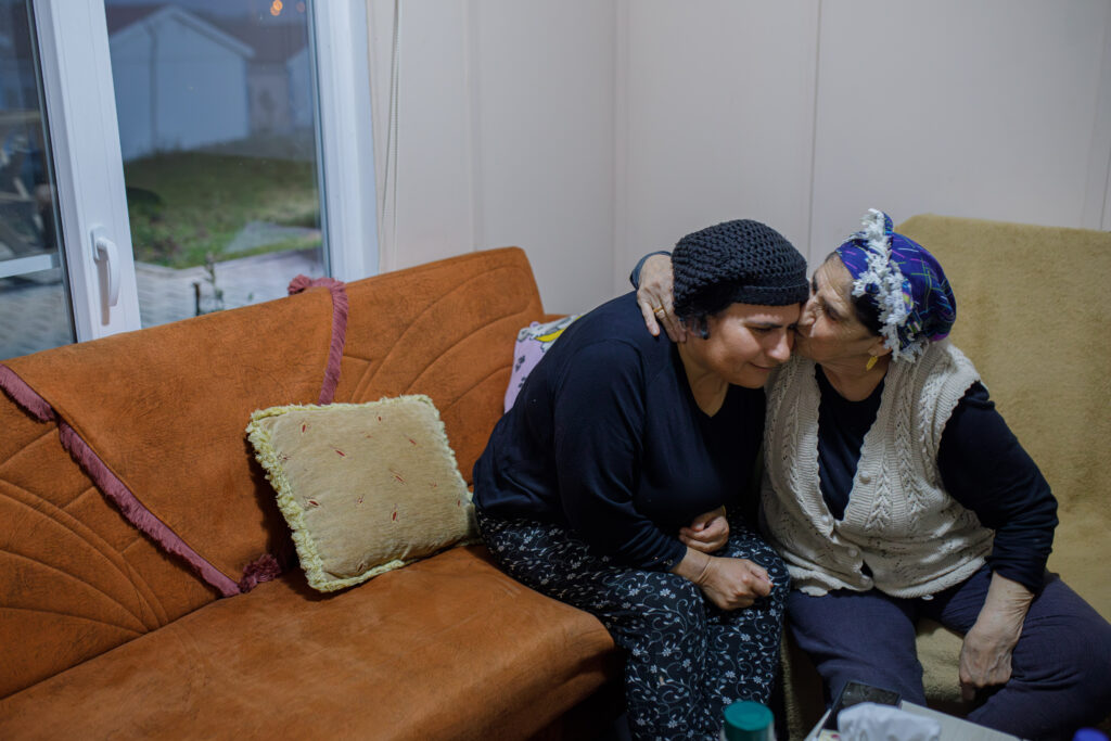 Saniye Yilmaz, who lives with her elderly parents in a prefabricated city in Hatay and has Parkinson's disease, sits on an orange sofa. Saniye Yilmaz's 76-year-old mother Sakine Yilmaz kisses her daughter on the forehead. Saniye is wearing a navy knit hat, long sleeved navy shirt, and floral print navy pants. Her mother is wearing a blue patterned scarf on her head, a beige sweater vest, and navy long-sleeved shirt and pants. Photo: Can Erok