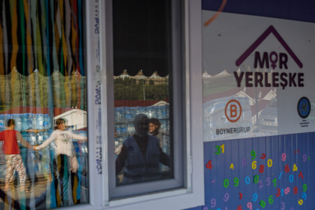 Children play in a prefabricated city west of Antakya. A psychologist and "social welfare officers" accompany the children as they play nursery rhymes outside one of the women's federation's "Purple Sites." You can see their reflection in the window, which has colorful paper streamers on the inside. Photo: Can Erok