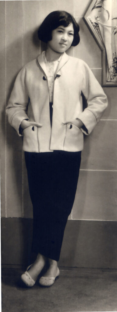 A photo of the author's grandmother, leaning against a wall. She's wearing a jacket with patch pockets, with both hands in her pockets. One leg is crossed over the other.