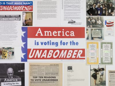 A photograph from the exhibition "Jokes About Bombs Will Not Be Taken Lightly" at the gallery Goswell Road, depicting the work of Lydia Eccles from her election campaign for the Unabomber in 1995-96. A bunch of paper ephemera on the wall, including photos, fliers, and bumper stickers. In the middle is a lawn sign that reads "America is voting for for the UNABOMBER".