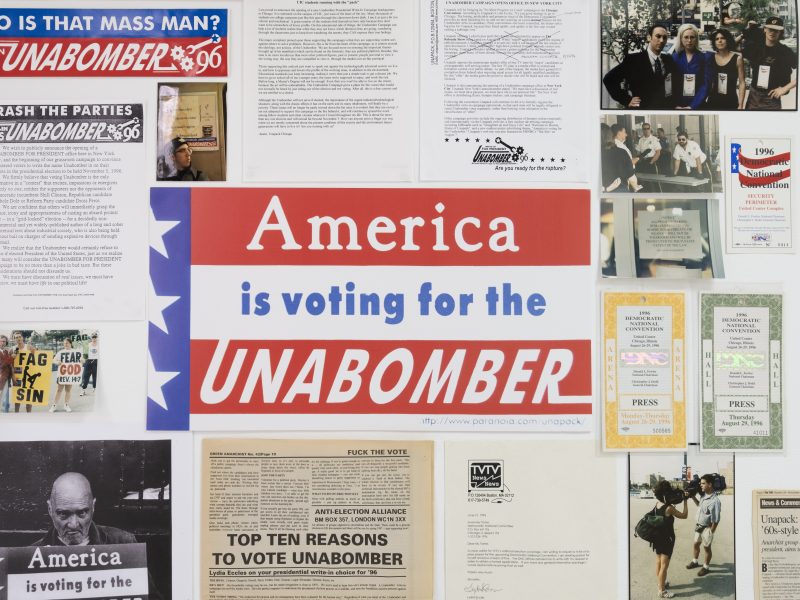 A photograph from the exhibition "Jokes About Bombs Will Not Be Taken Lightly" at the gallery Goswell Road, depicting the work of Lydia Eccles from her election campaign for the Unabomber in 1995-96. A bunch of paper ephemera on the wall, including photos, fliers, and bumper stickers. In the middle is a lawn sign that reads "America is voting for for the UNABOMBER".