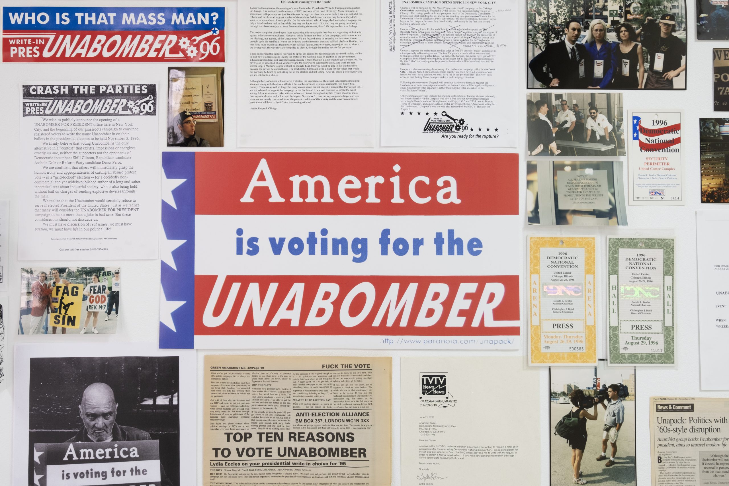 A photograph from the exhibition "Jokes About Bombs Will Not Be Taken Lightly" at the gallery Goswell Road, depicting the work of Lydia Eccles from her election campaign for the Unabomber in 1995-96. A bunch of paper ephemera on the wall, including photos, fliers, and bumper stickers. In the middle is a lawn sign that reads "America is voting for for the UNABOMBER".