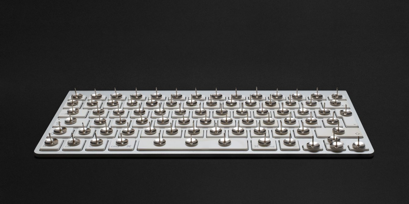 A modern white keyboard on a black background. On each key, there's a thumbtack with the sharp end pointing upwards.
