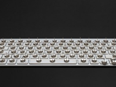 A modern white keyboard on a black background. On each key, there's a thumbtack with the sharp end pointing upwards.