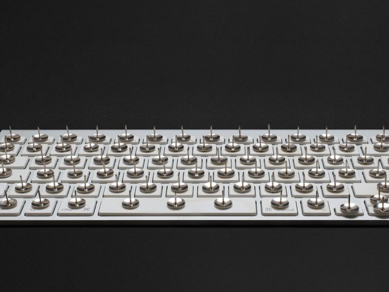 A modern white keyboard on a black background. On each key, there's a thumbtack with the sharp end pointing upwards.