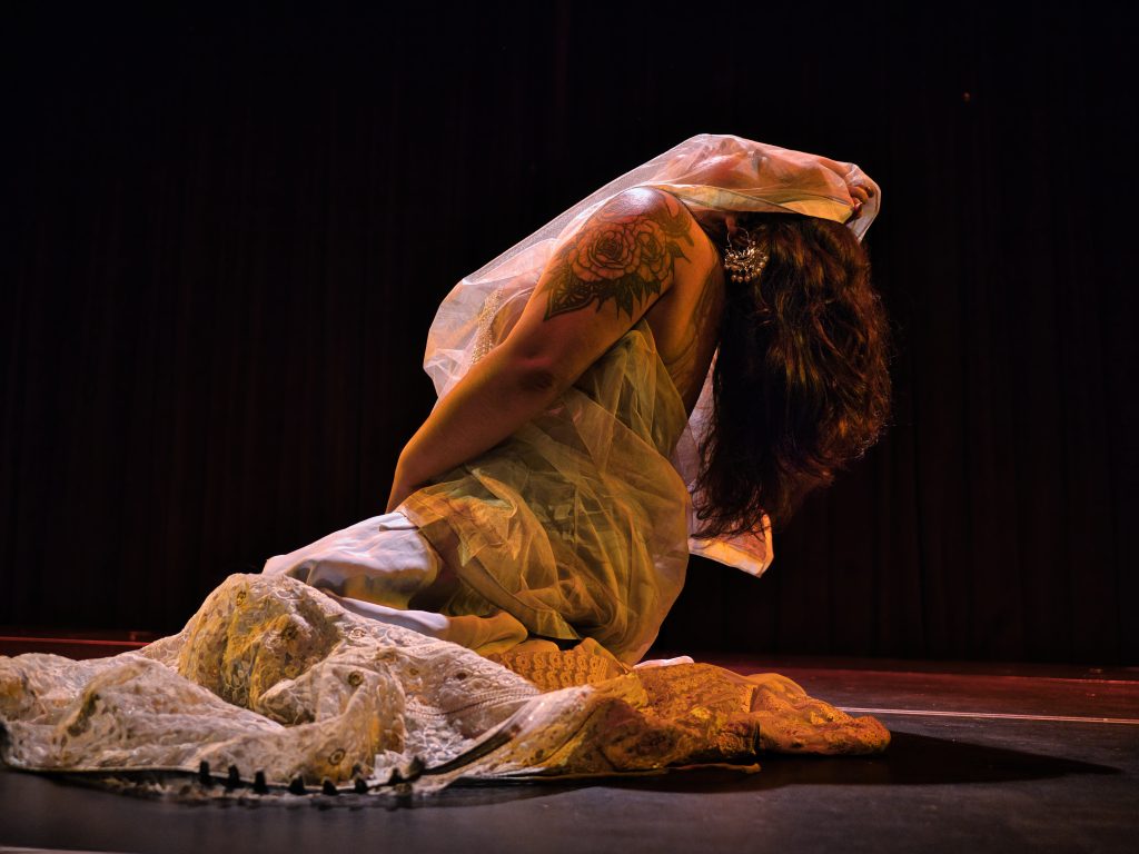 A photo of Amita Vempati, mid-performance. She is in profile, on her knees, with her head thrown back; wrapped in fabric, a sheer panel covering her face.