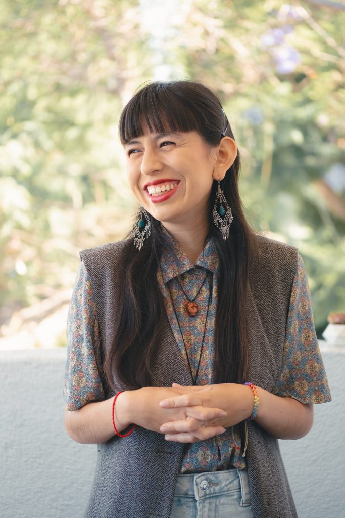 A portrait photo of Anaís Azul, smiling and looking off-camera. They have long, dark brown hair with bangs, and are wearing red lipstick, dangly earrings, and a gray vest over a patterned shirt. Their hands are crossed in front of their body, fingers intertwined. 