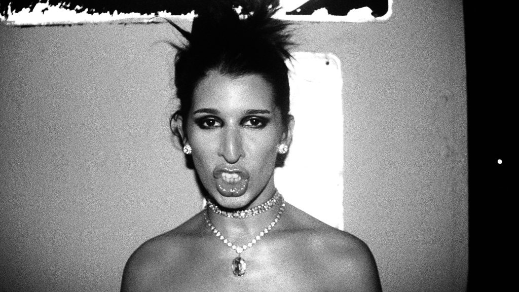 A black and white photo of AV María, seemingly mid-word. Her hair is tied back in a high, messy bun; she's wearing earrings, a choker, and a slightly longer necklace.