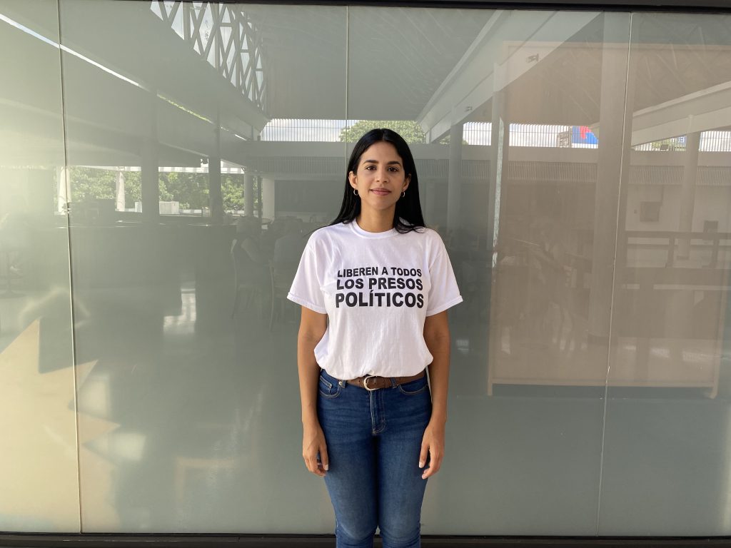 A photo of Sairam Rivas standing in front of a glass wall. She is wearing a white t-shirt with black text that reads, "Liberen a todos los presos políticos."