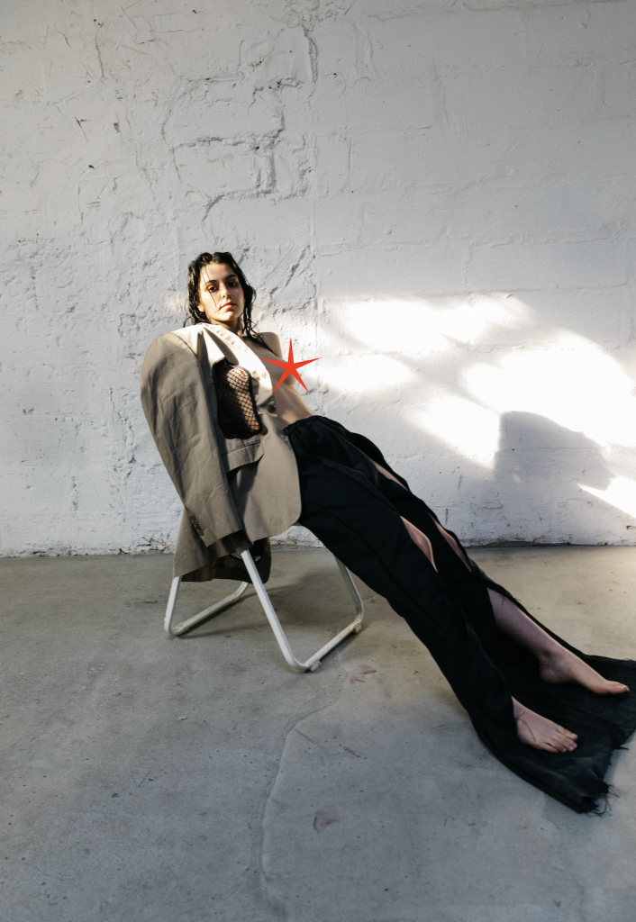 A portrait of slic, sitting in a folding chair in front of a white brick wall. They have a greige blazer with a fishnet insert draped over one shoulder, and are wearing what appears to be a long skirt/pants with slits throughout. They're looking at the camera.