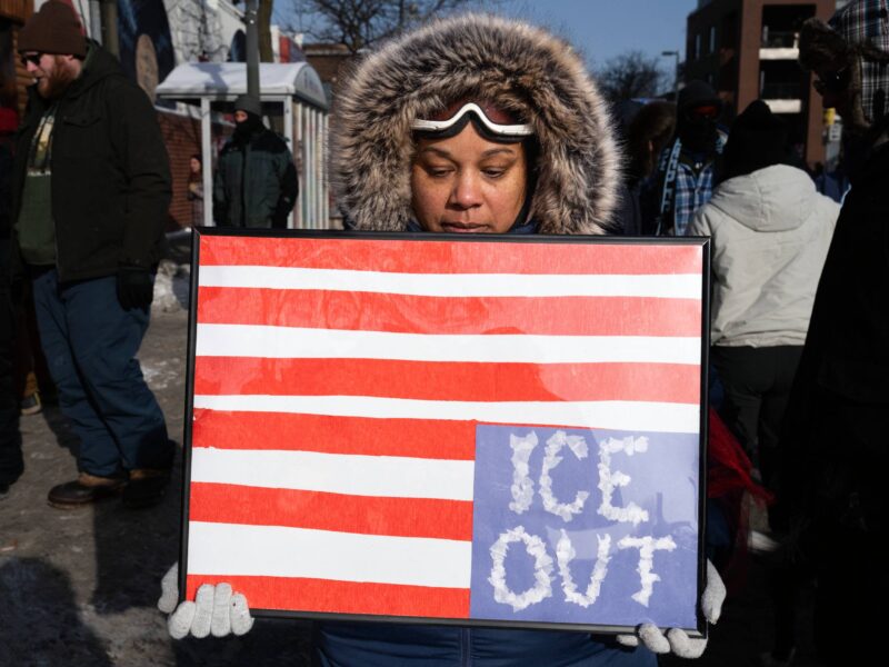 A protestor in a coat holding a sign designed like an upside down American flag, with "ICE OUT" written in the blue section.