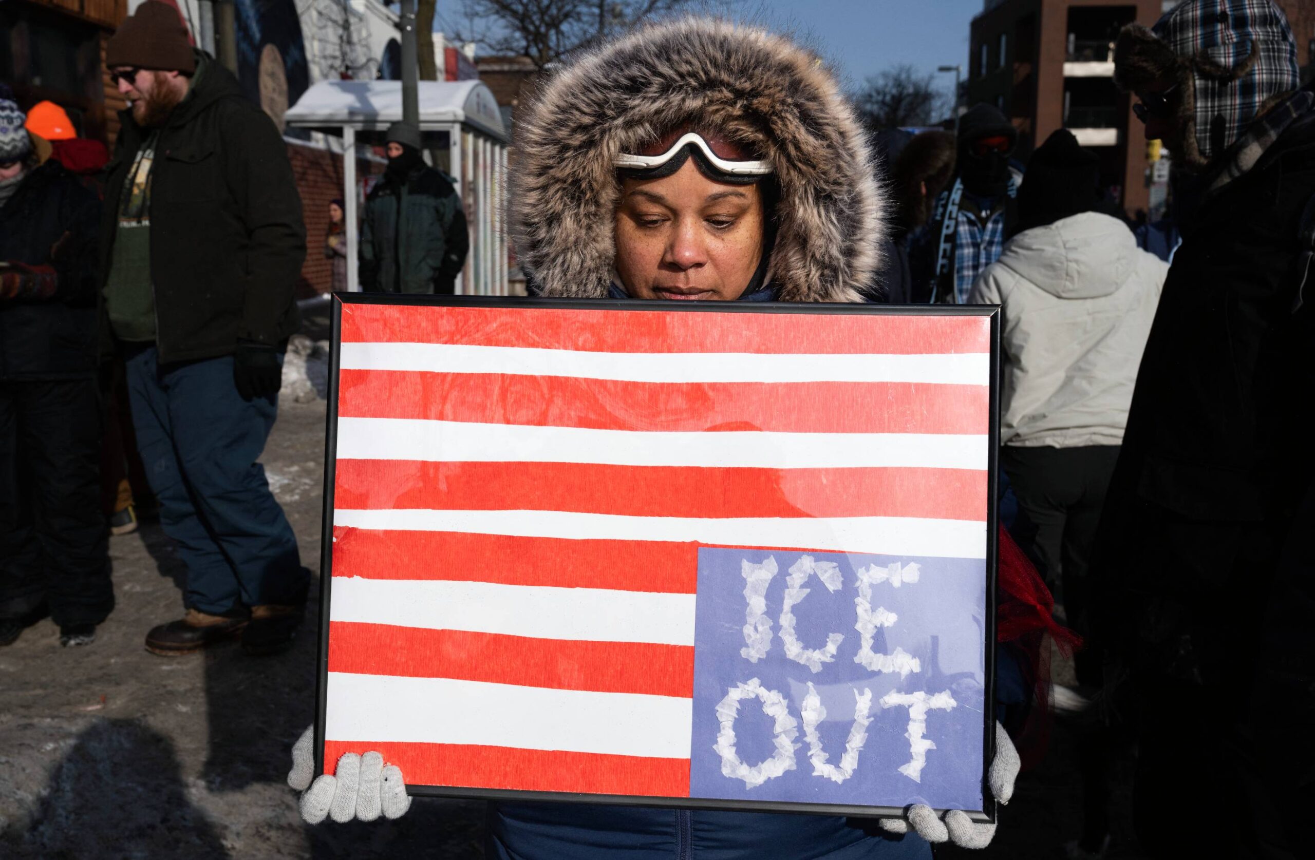 A protestor in a coat holding a sign designed like an upside down American flag, with "ICE OUT" written in the blue section.